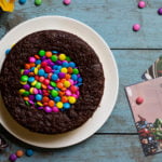 A colorful M&M Pinata Cake topped with chocolate ganache